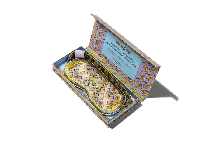 A colorful slip silk sleep mask with a zodiac Aquarius design, featuring yellow and purple tones. Displayed in branded open packaging box on white background.