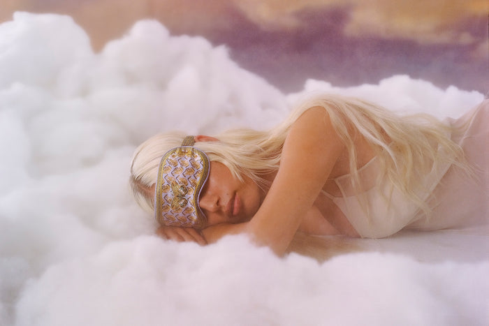 A woman wearing a colorful slip silk sleep mask with a zodiac Aquarius design, featuring yellow and purple tones, laying in the clouds.