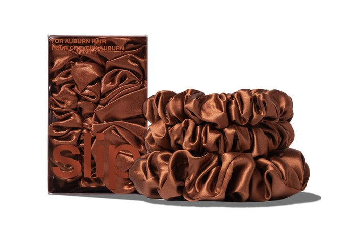A set of three auburn colored slipsilk scrunchies, including two midi and one large, stacked in front of branded packaging on a white background.