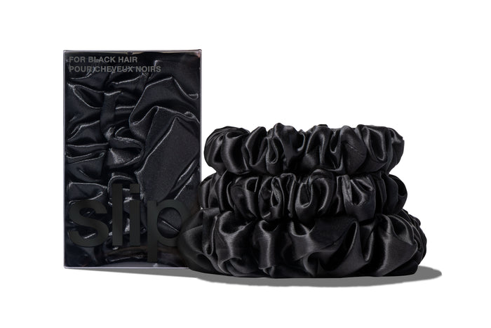 A set of three black colored slipsilk scrunchies, including two midi and one large, stacked in front of branded packaging on a white background.