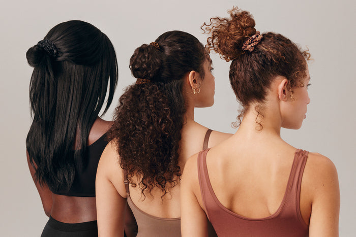 Three women with styled hair wearing slip silk skinny scrunchies in their hair in black, dark brown and light brown.