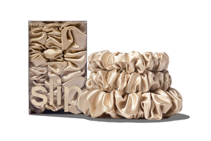 A set of three blonde colored slipsilk scrunchies, including two midi and one large, stacked in front of branded packaging on a white background.
