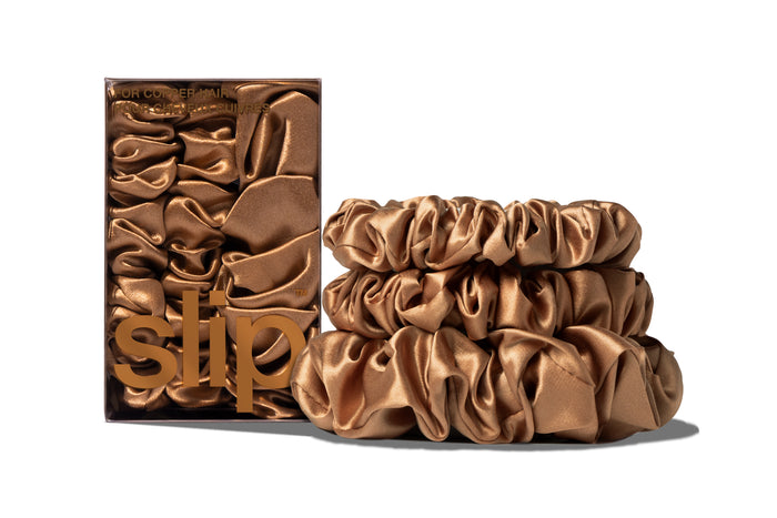 A set of three copper colored slipsilk scrunchies, including two midi and one large, stacked in front of branded packaging on a white background.