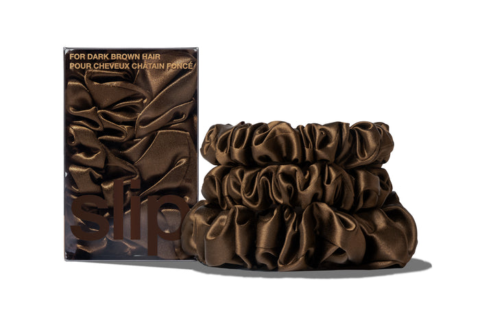 A set of three dark brown colored slipsilk scrunchies, including two midi and one large, stacked in front of branded packaging on a white background.