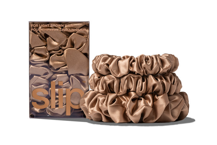 A set of three light brown colored slipsilk scrunchies, including two midi and one large, stacked in front of branded packaging on a white background.