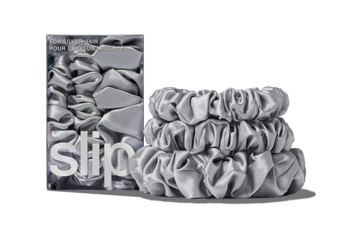 A set of three silver colored slipsilk scrunchies, including two midi and one large, stacked in front of branded packaging on a white background.