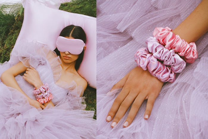 Woman lying down on a lilac slip silk queen pillowcase in a lilac dress, wearing a slip lilac contour mask and large scrunchies. Hand with three large pink and  lilac slip scrunchies.