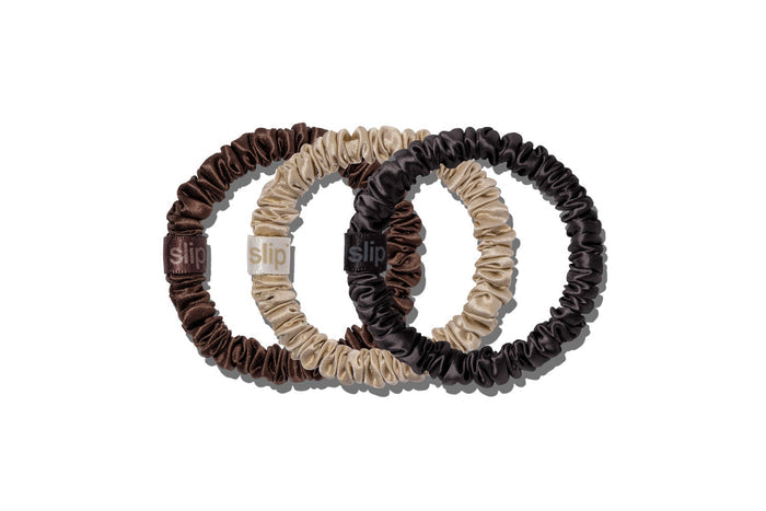 Three pure silk hair scrunchies in dark brown, beige, and black, overlapping on a clean white background. Each scrunchie features a small fabric brand tag with the word 'slip'.