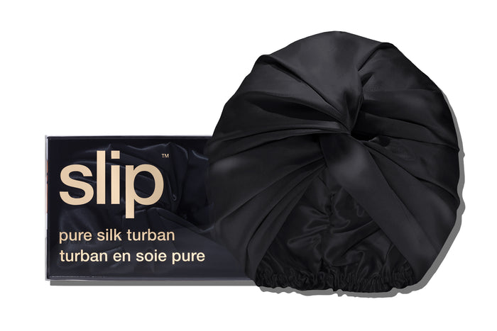 A black slip silk turban in front of with branded packaging on a white background.