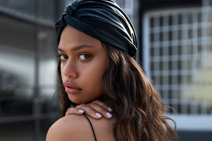 A woman wearing black slip silk turban with blurred architectural background.