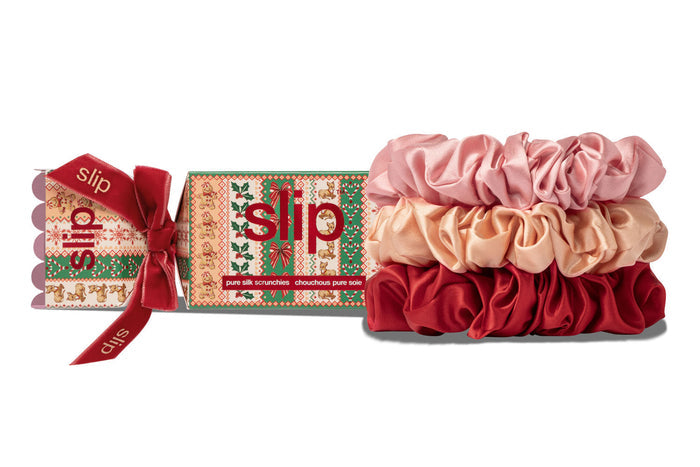 Three slip silk hair scrunchies in pink, peach, and red are displayed next to their Christmas-themed packaging box with a red ribbon, on a white background.