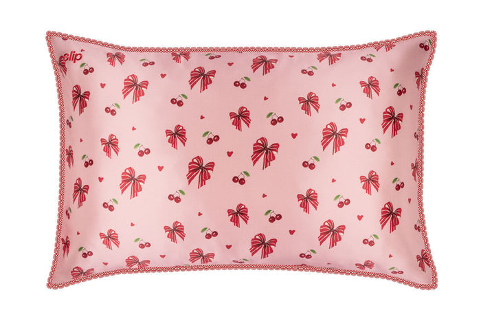 A slip silk pillowcase featuring a repeating pattern of red bows, cherries, and small hearts, finished with a scalloped lace trim.