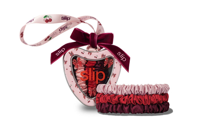 A heart-shaped Slip scrunchie gift set containing three silk scrunchies in pink, red, and burgundy, packaged in a heart-shaped tin box with a dark burgundy ribbon and a matching printed hanging ribbon.