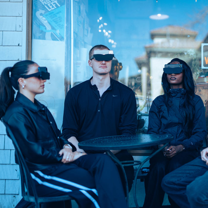 Three people wearing futuristic goggles sitting around a table in an urban setting.