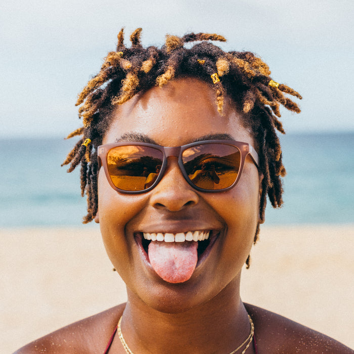 Person with dreadlocks wearing sunglasses and sticking out their tongue on a beach.