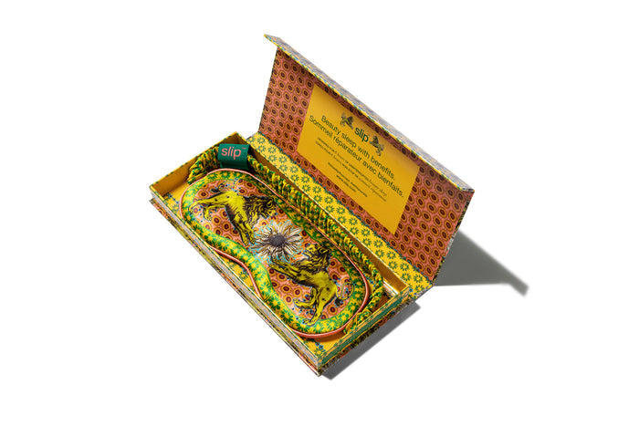 A colorful slip silk sleep mask with a zodiac Leo design, featuring yellow and gold tones. Displayed in branded open packaging box on white background.