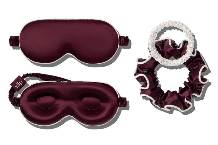 A sleep and haircare set featuring a crimson contour sleep mask with white piping, and matching large silk scrunchie, and a white skinny scrunchie, arranged on a white background.