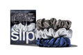 A set of three slip silk large scrunchies in navy, silver, and charcoal, displayed in front of branded packaging on a white background.