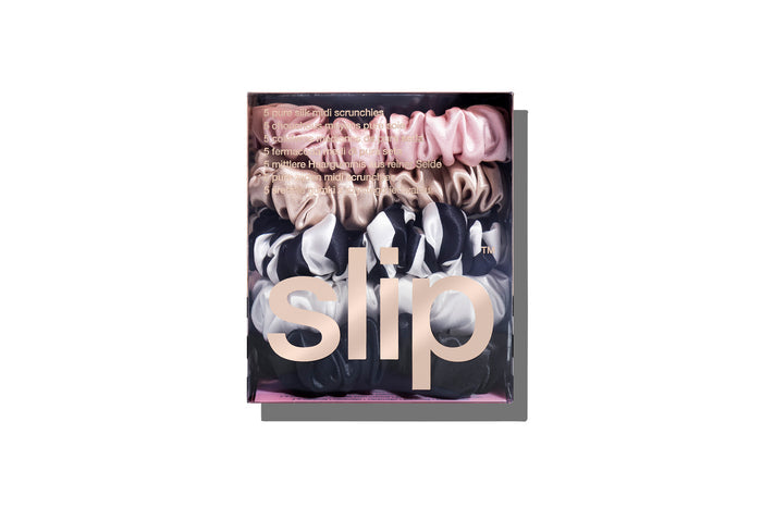 A set of five Slip silk midi scrunchies in various colors including black, pink, caramel, white, and navy stripe in branded packaging on a white background.