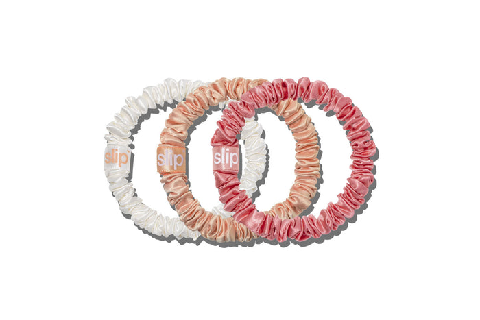 Three slip silk skinny hair scrunchies in white, peach, and pink, arranged in a circular overlapping pattern on a solid white background. The brand name 'slip' is visible on the tag of each scrunchie.