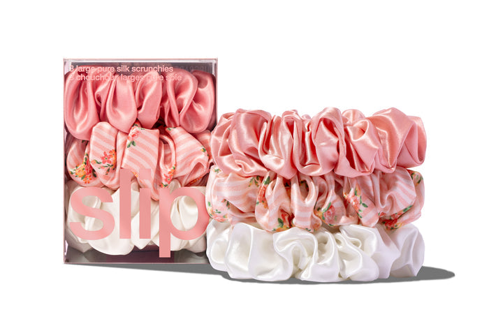 Stack of pink, floral pattern and white large slip silk scrunchies, displayed in front of branded packaging on a white background.