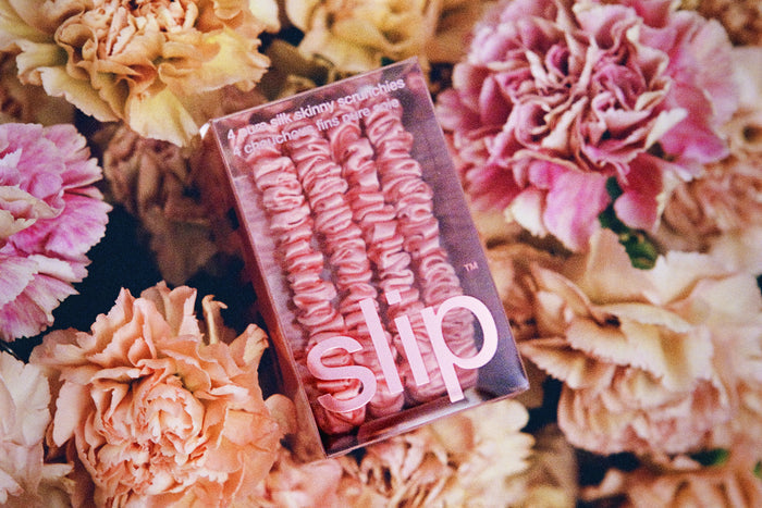 Blush pink silk skinny scrunchies in slip packaging on a bed of flowers.