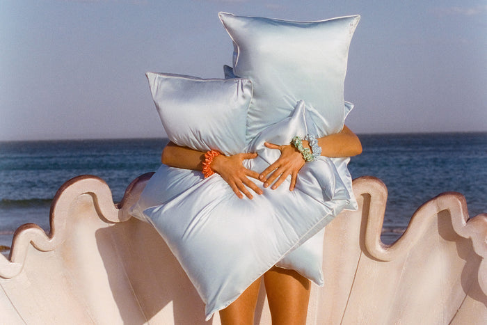 Model holding many light blue silk pillowcases in front of the ocean.